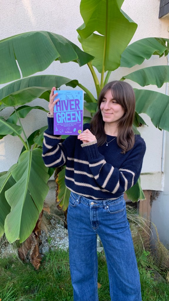 Une femme tient un livre intitulé Hiver Green dans les mains en extérieur devant un bananier.